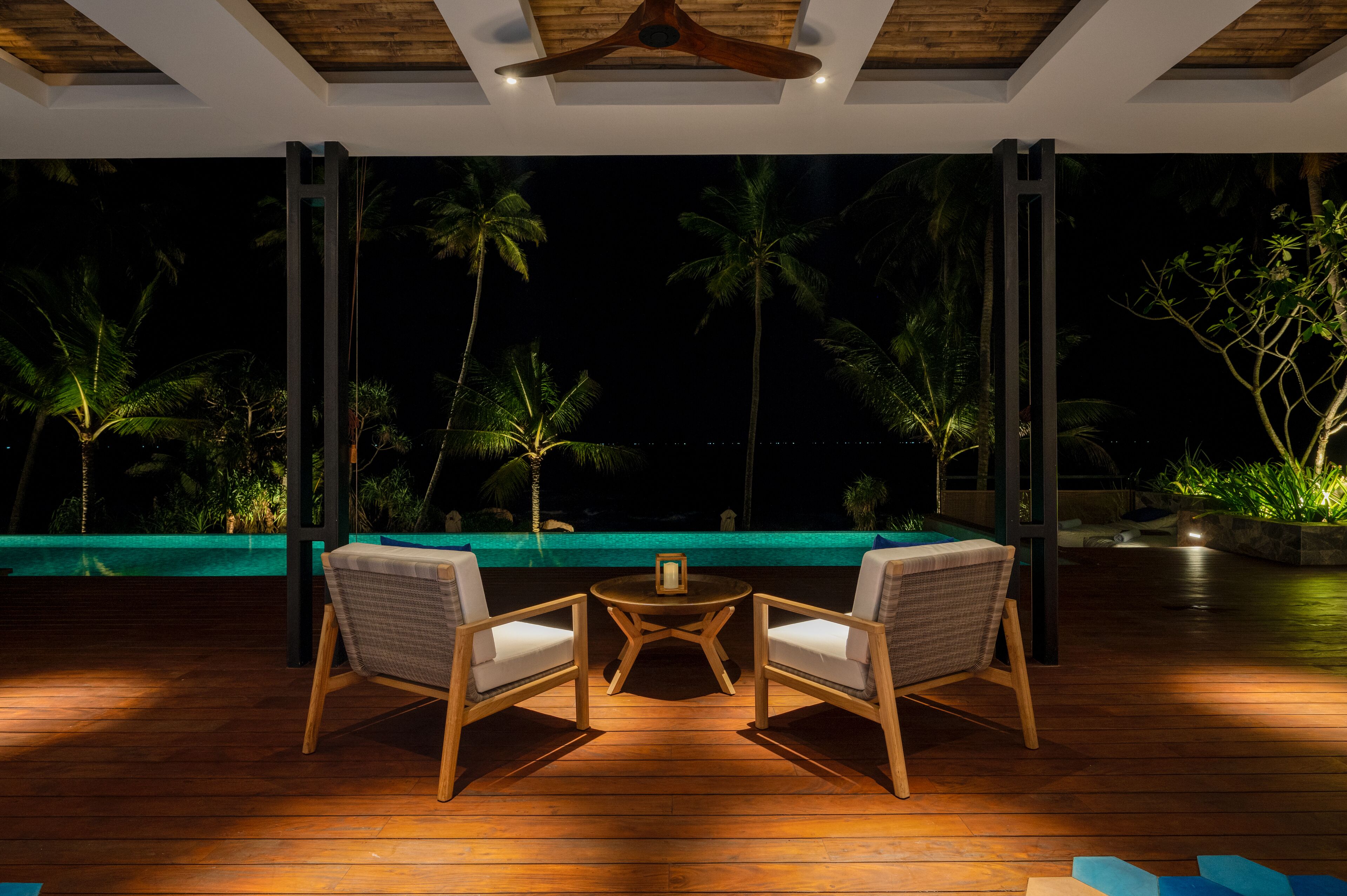 An outdoor seating area with two chairs and a table, overlooking a pool with palm trees and a night sky.