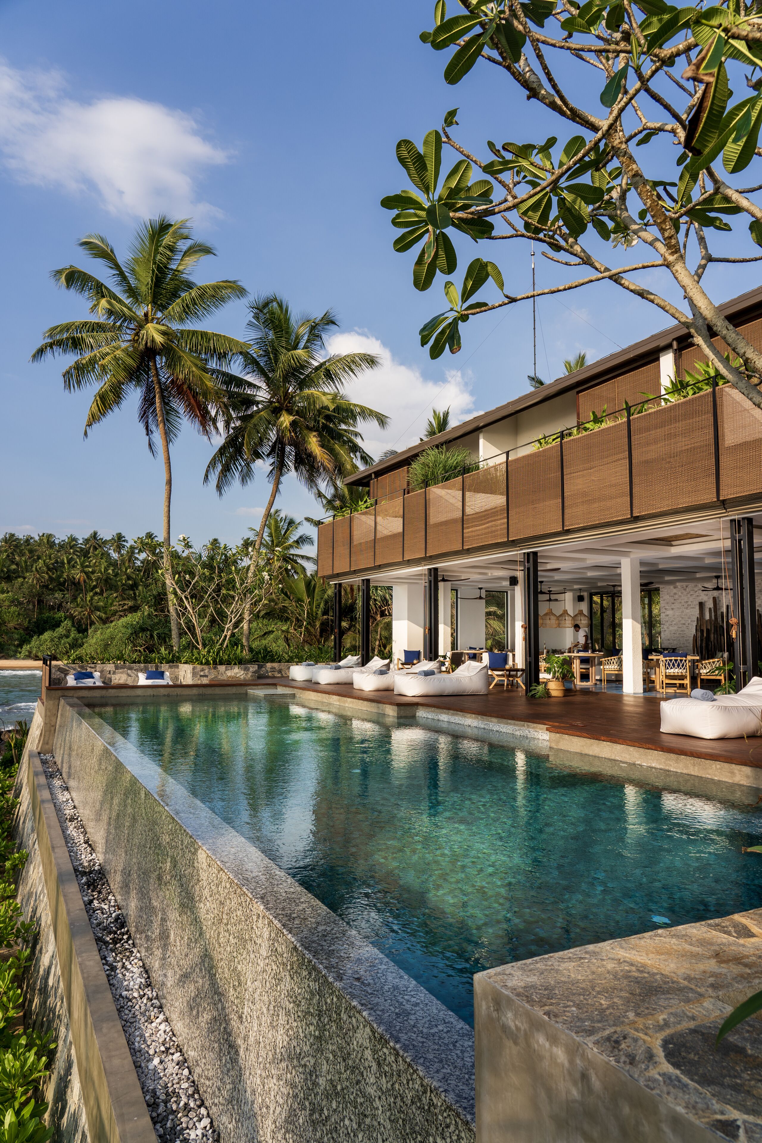 A modern poolside area with a clear pool, lounge chairs, and a covered seating area with tables.