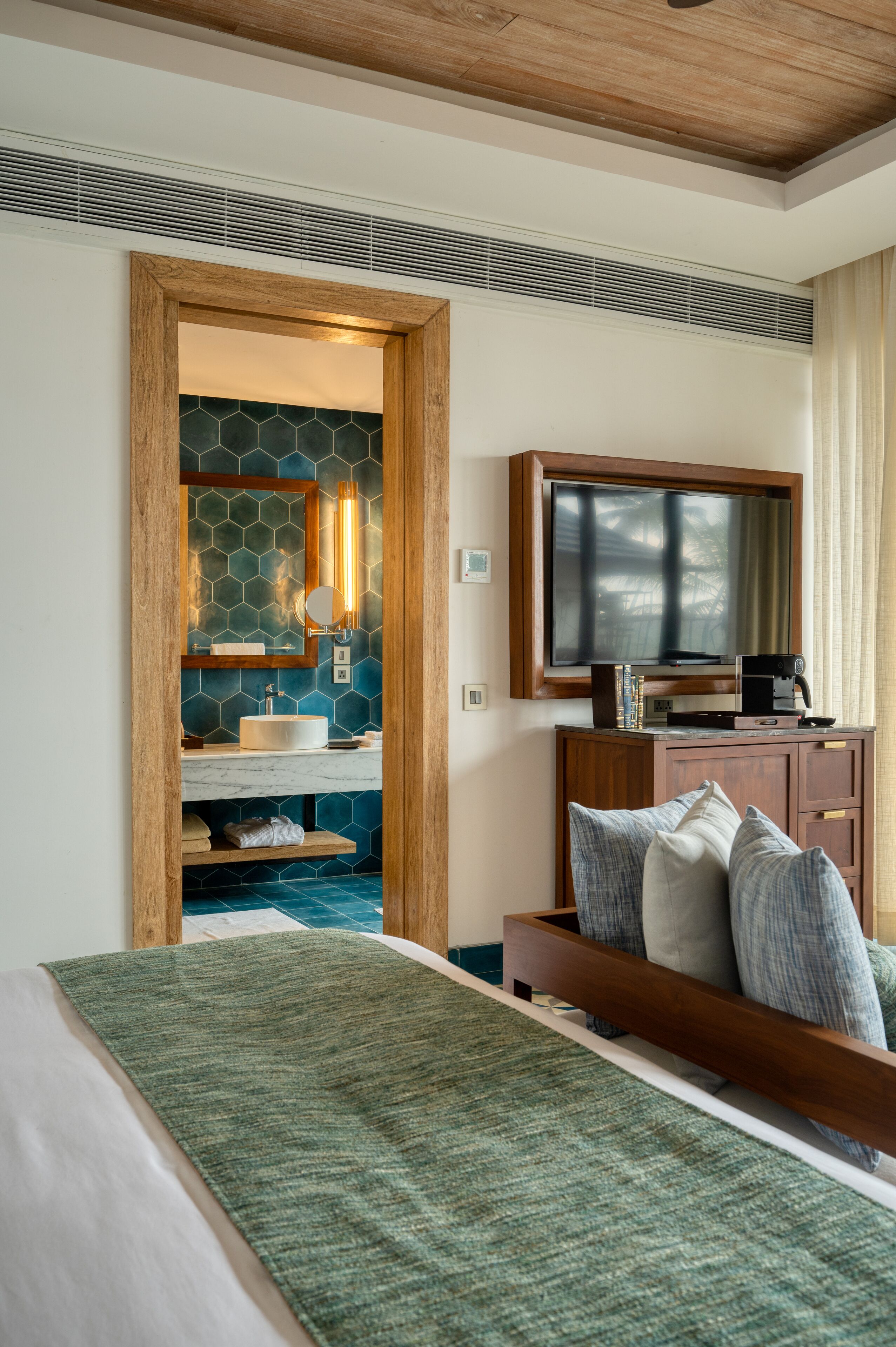 A hotel room with a bed, a wooden headboard, a television, a wooden cabinet, and a bathroom with a mirror, sink, and tiled backsplash.