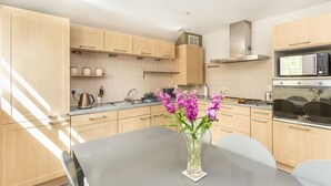 A modern kitchen with light wood cabinets, a stainless steel microwave, and a vase with purple flowers on the table. - An Sulaire (North Berwick)
