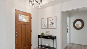Un pasillo con una puerta de madera, una consola negra con dos impresiones enmarcadas de motivos botánicos, una planta pequeña en maceta y un espejo redondo en la pared.