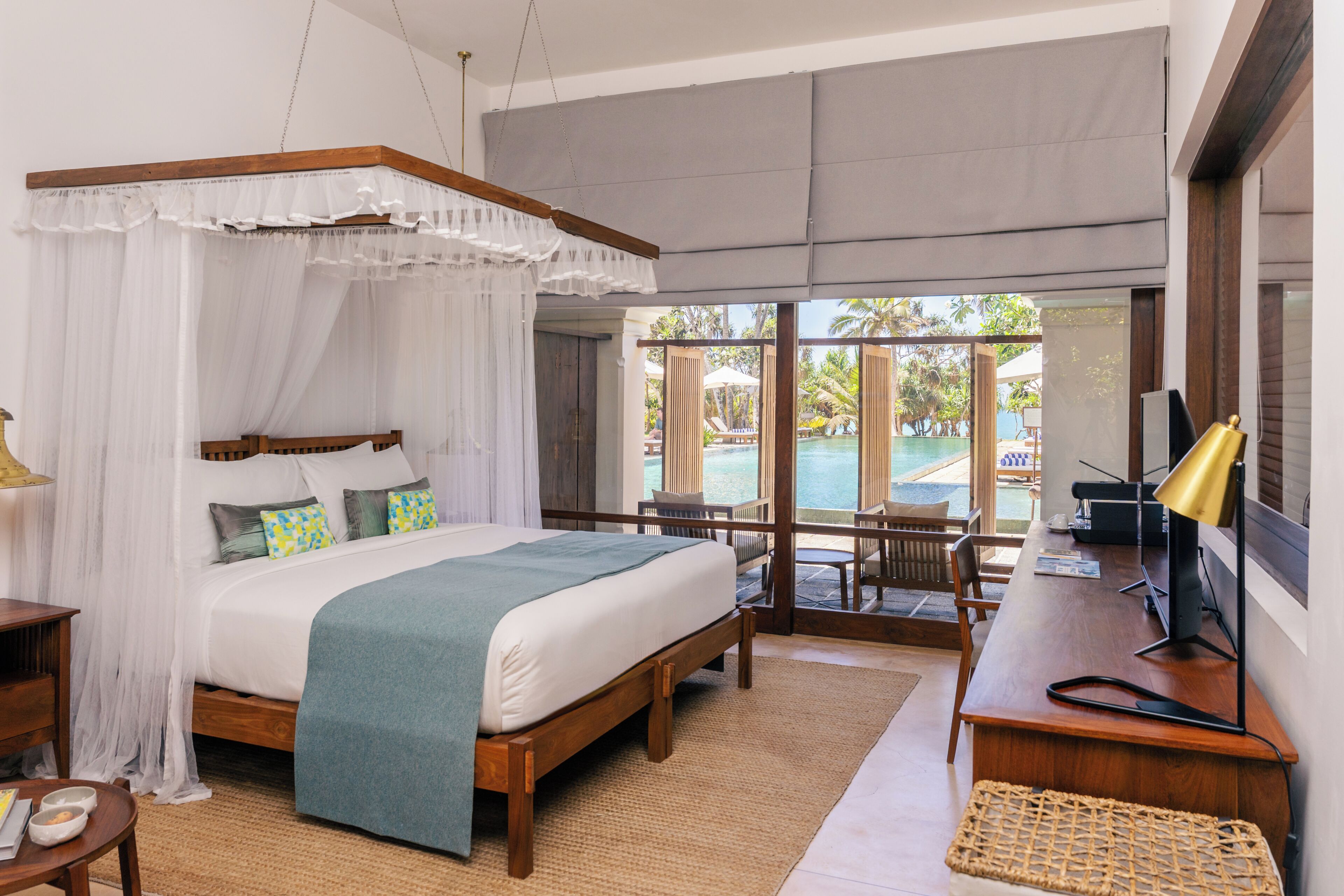 A bedroom with a four-poster bed, a desk with a lamp, and a view of an outdoor pool area.