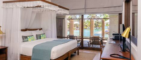 A bedroom with a four-poster bed, a desk with a lamp, and a view of an outdoor pool area.