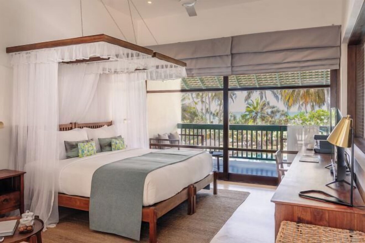 A bedroom with a bed, a desk, a chair, and a balcony with a view of palm trees.