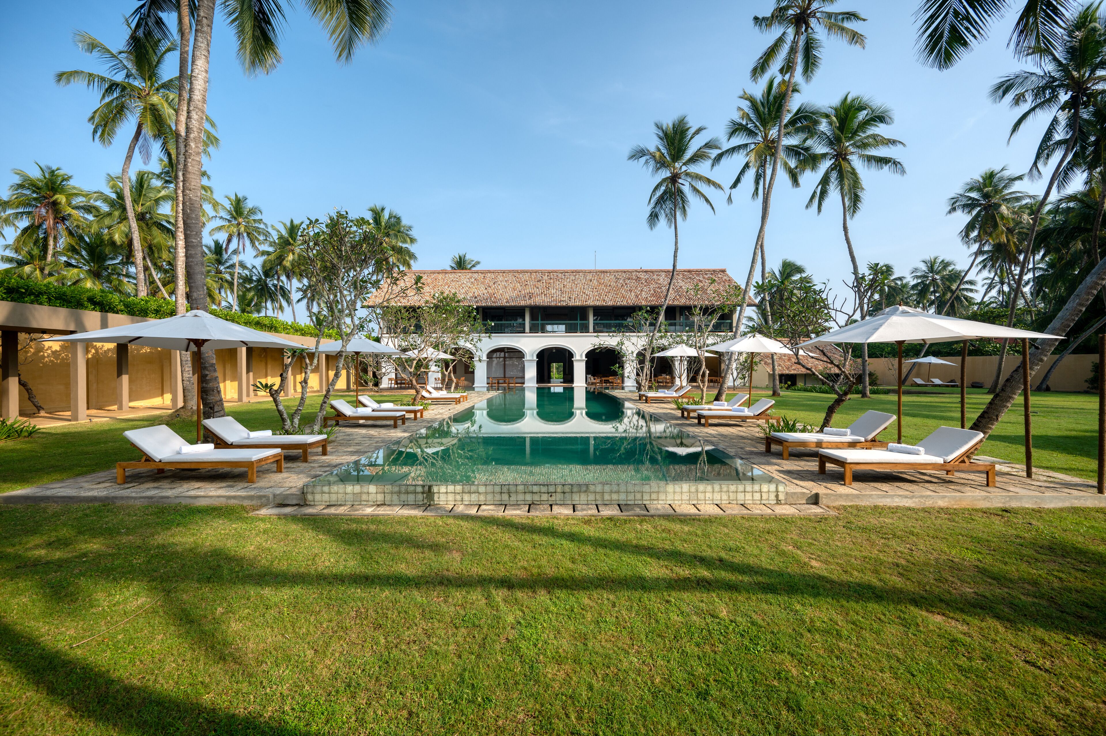 A pool area with lounge chairs and umbrellas, surrounded by palm trees and a building with a tiled roof.