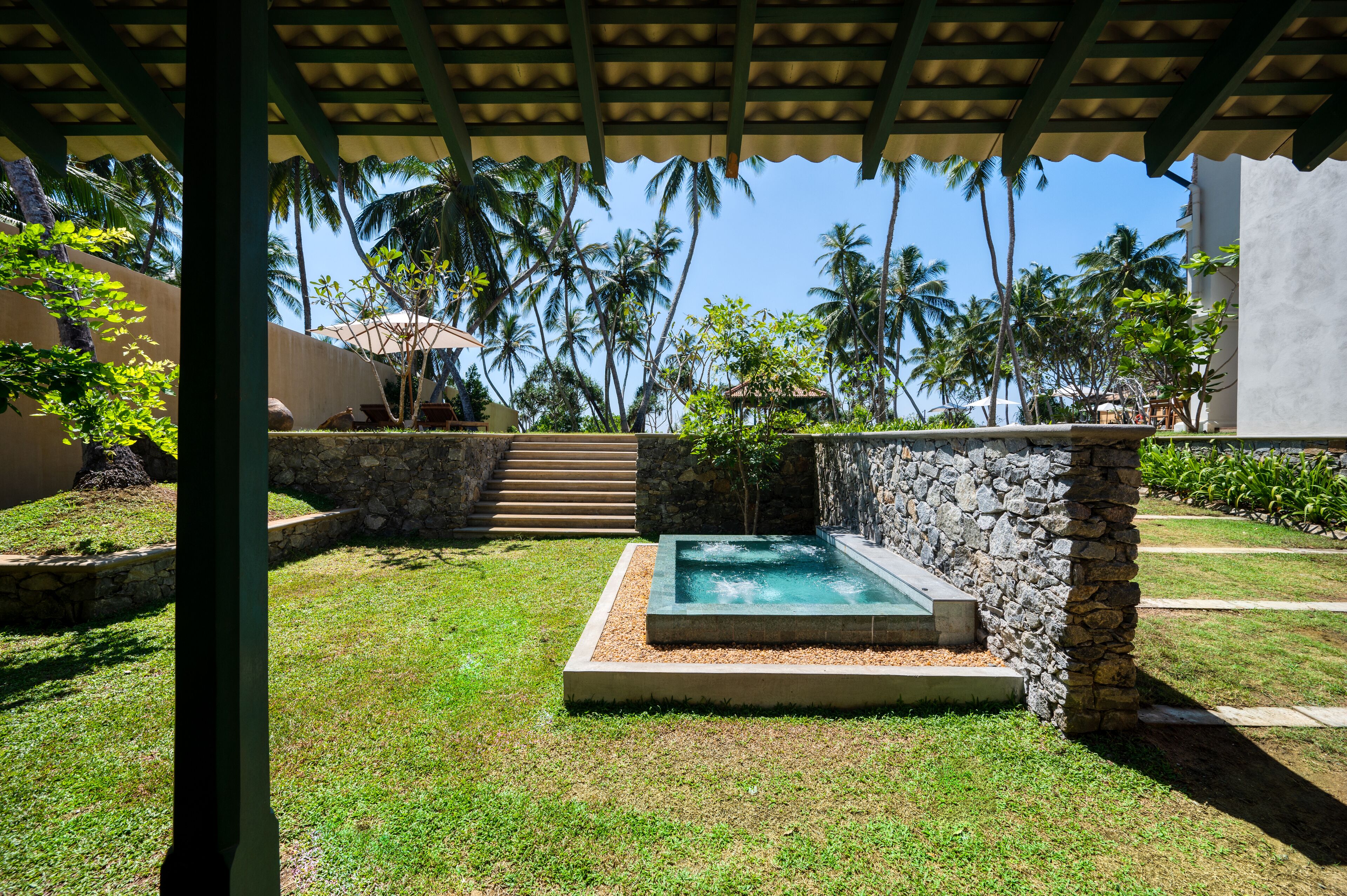 Un espacio al aire libre techado con un muro de piedra, una pequeña piscina y una escalera que sube a un nivel superior.