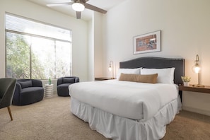 A bedroom with a large bed, two bedside tables, a ceiling fan, a window with blinds, and a framed picture on the wall.