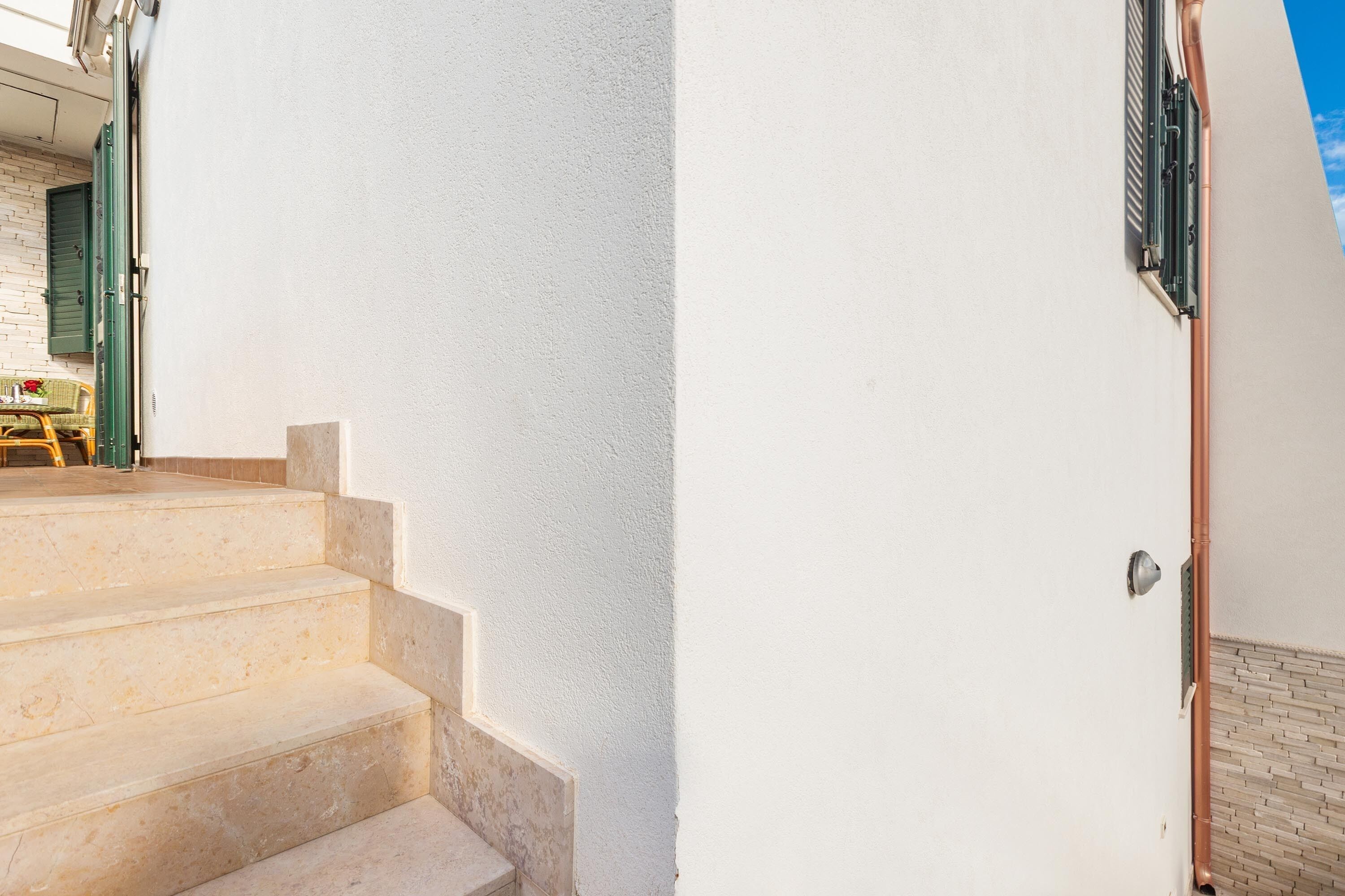 A set of beige stone steps leading to a white door with a green shutter, a copper pipe, and a small window with a metal shutter.