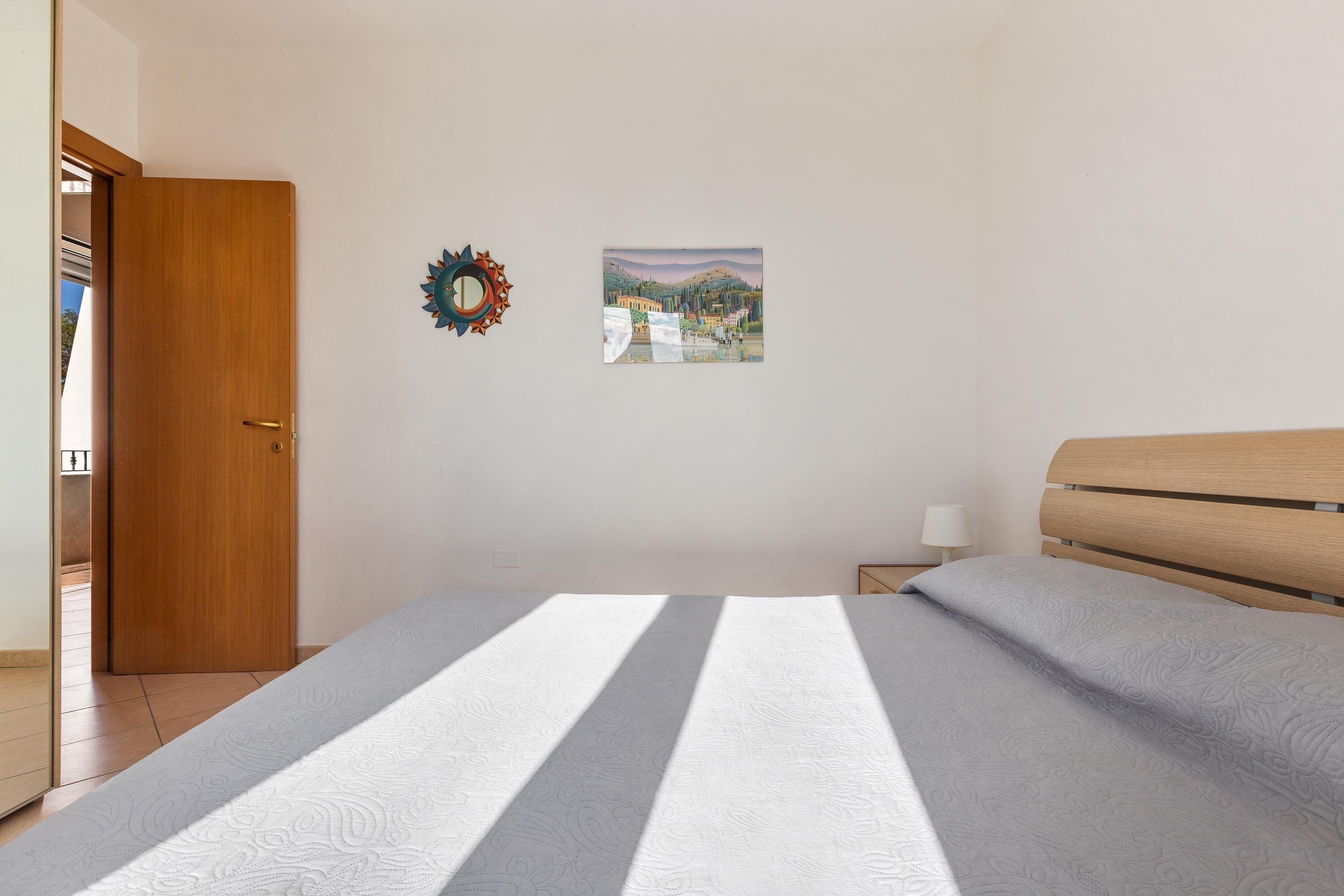 A bedroom with a bed, a wooden door, a small bedside table with a lamp, and a wall decoration.