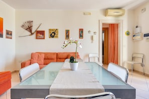 A dining area with a glass table, orange sofa, and a potted orchid.