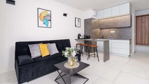 Una sala de estar moderna con un sofá negro, una mesa de centro con un florero, y un área de cocina con un taburete de bar y armarios.