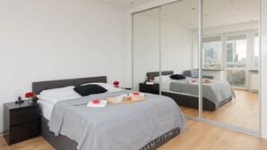 Un dormitorio moderno con un espejo grande, una cama con ropa de cama gris, una mesita de noche con una lámpara y una bandeja con artículos para el desayuno.