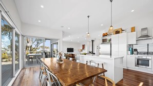 A modern kitchen with white cabinetry, a wooden dining table, and a large window overlooking a garden. - Beach Comber (Anglesea)