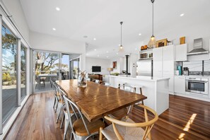 A modern kitchen with white cabinetry, a wooden dining table, and a large window overlooking a garden. - Beach Comber (Anglesea)