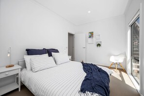 A modern bedroom with a bed, bedside tables, a nightstand, a lamp, a chair, and a window. - Beach Comber (Anglesea)