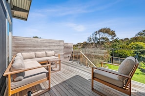 A wooden deck with a sofa and chairs, a glass balustrade, and a view of the surrounding greenery. - Beach Comber (Anglesea)