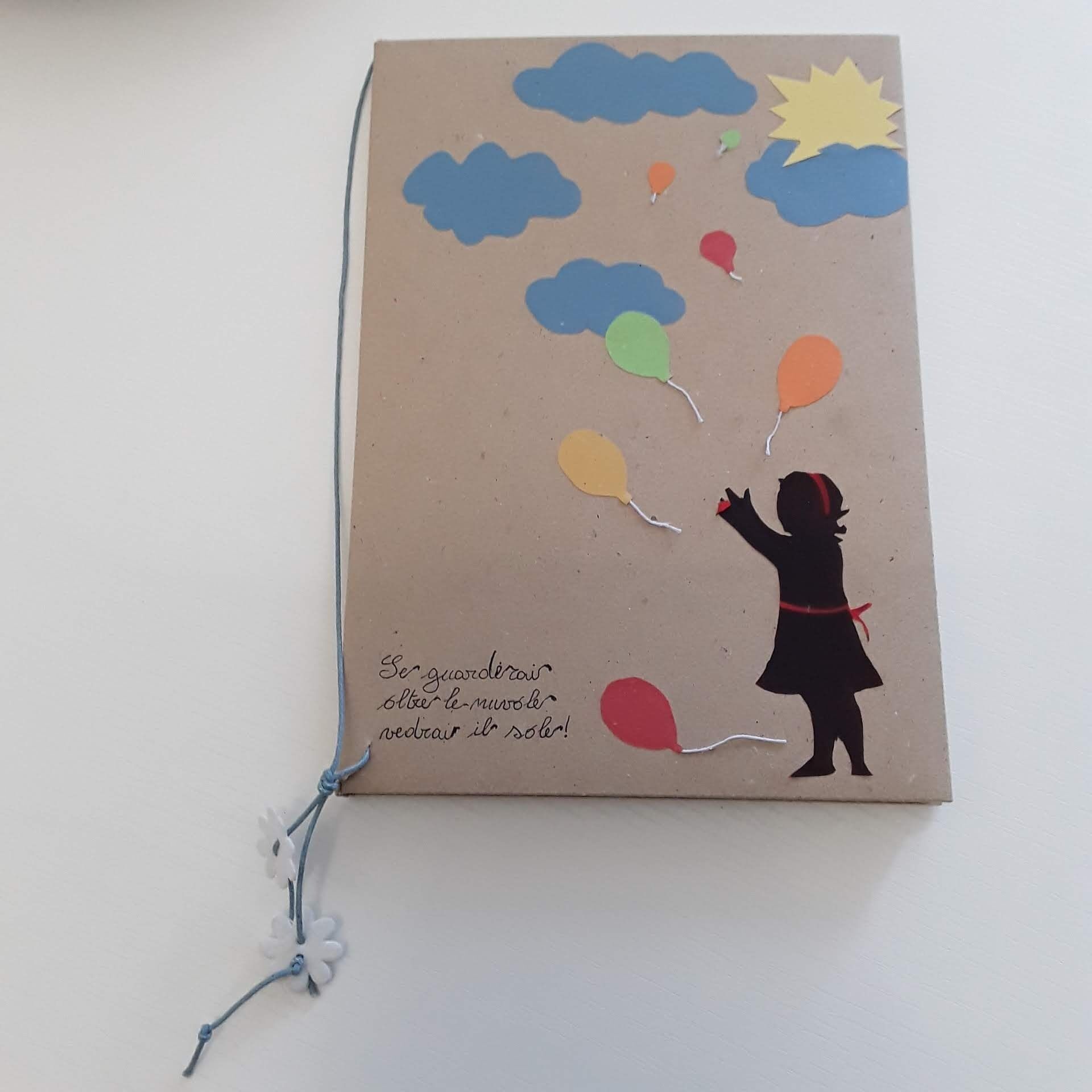 Ein Notizbuch mit einer Silhouette eines Kindes, das Ballons und Wolken hält, und einer handschriftlichen Botschaft in einer Fremdsprache.