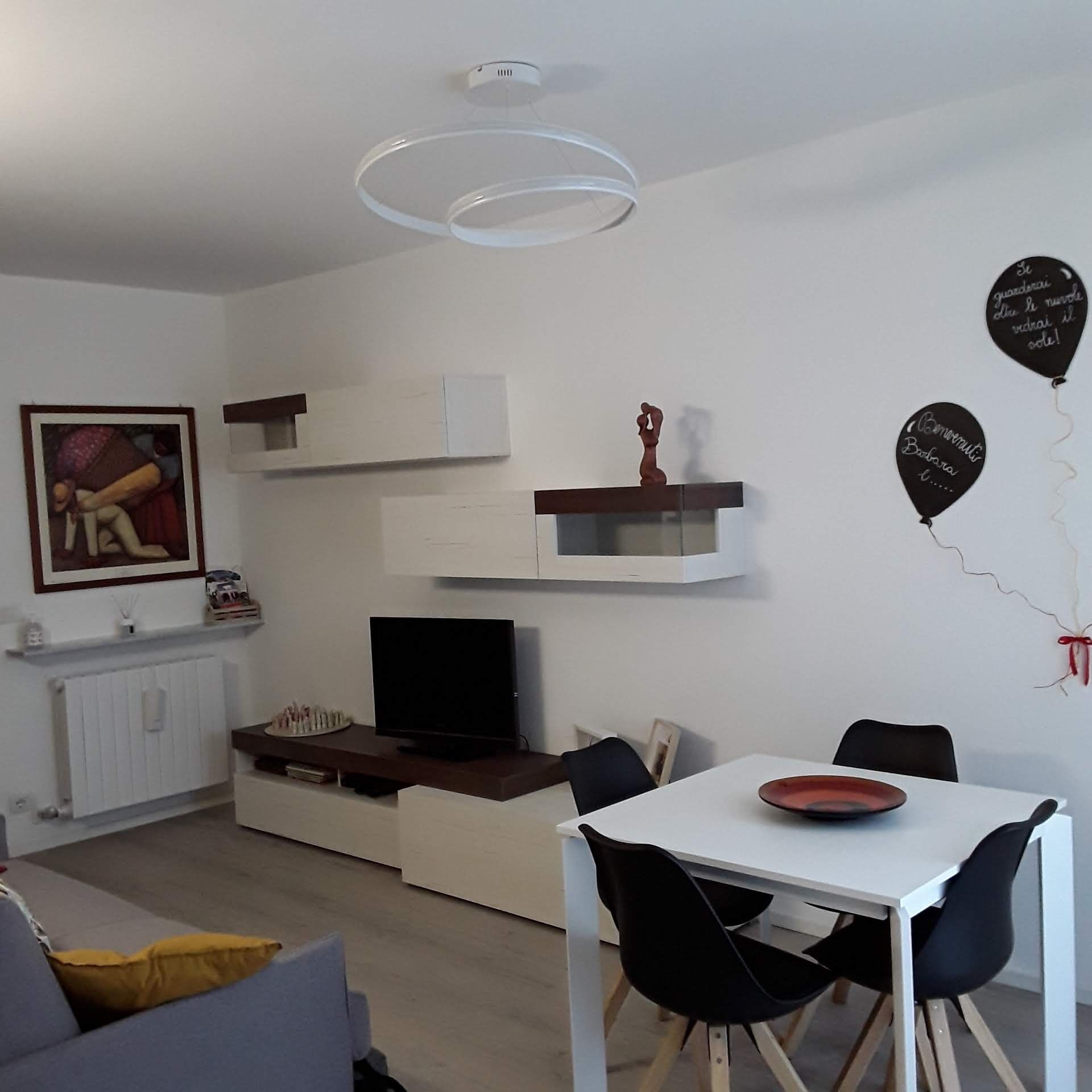 A modern living room with a flat-screen TV, a dining table with chairs, and a white ceiling light fixture.