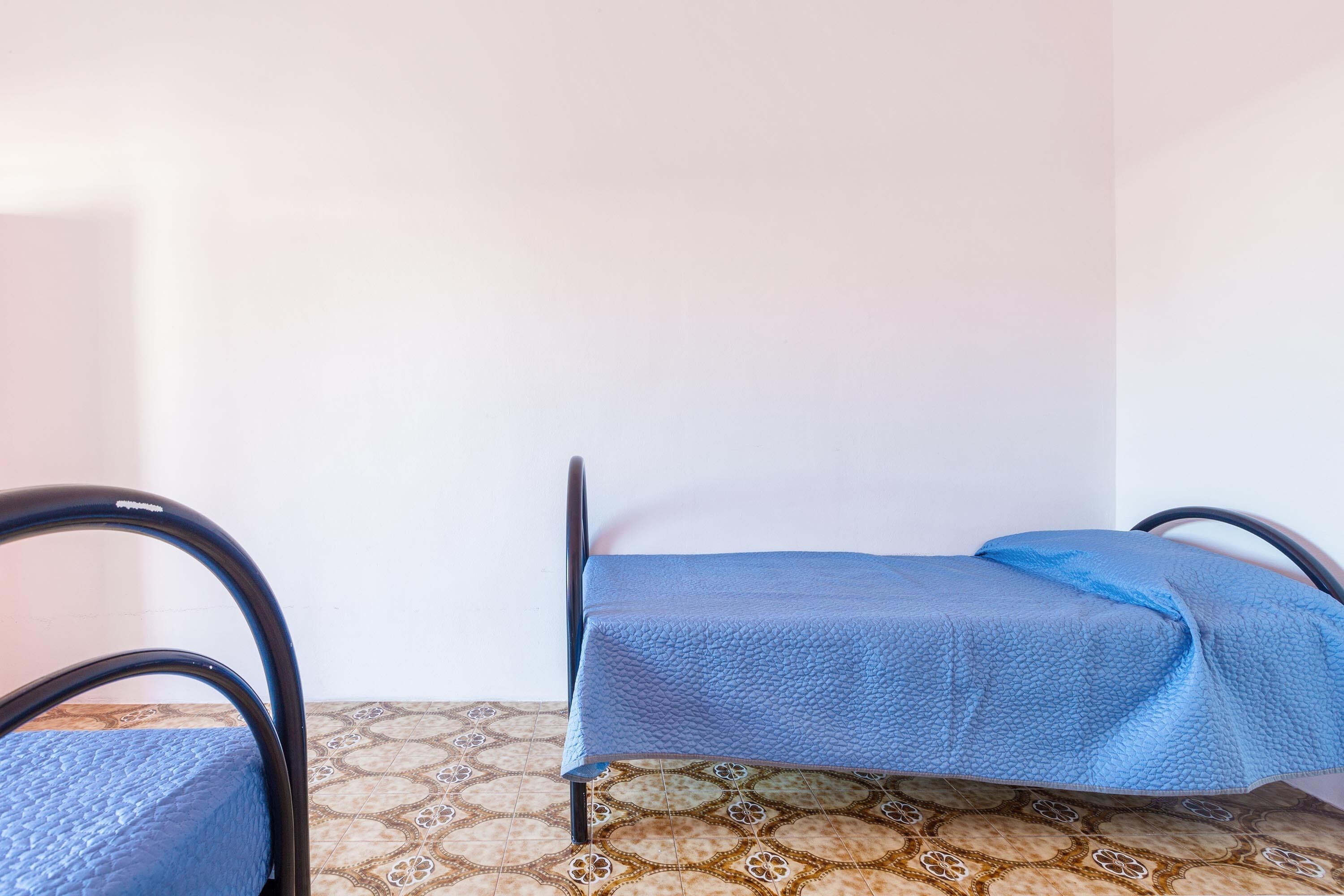A single bed with a blue bedspread in a room with patterned floor tiles and a plain wall.