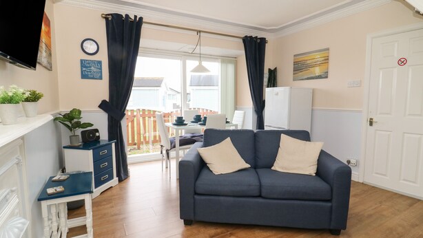Een woonkamer met een bank, een eettafel en een televisie. Er is een raam met gordijnen, een klok aan de muur en een deur met een bordje.