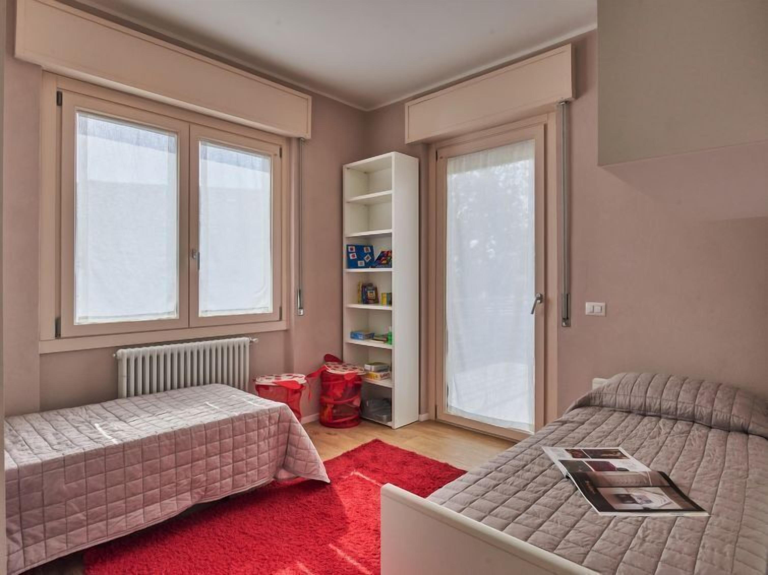 Une chambre avec deux lits, un tapis rouge, une bibliothèque et une fenêtre.