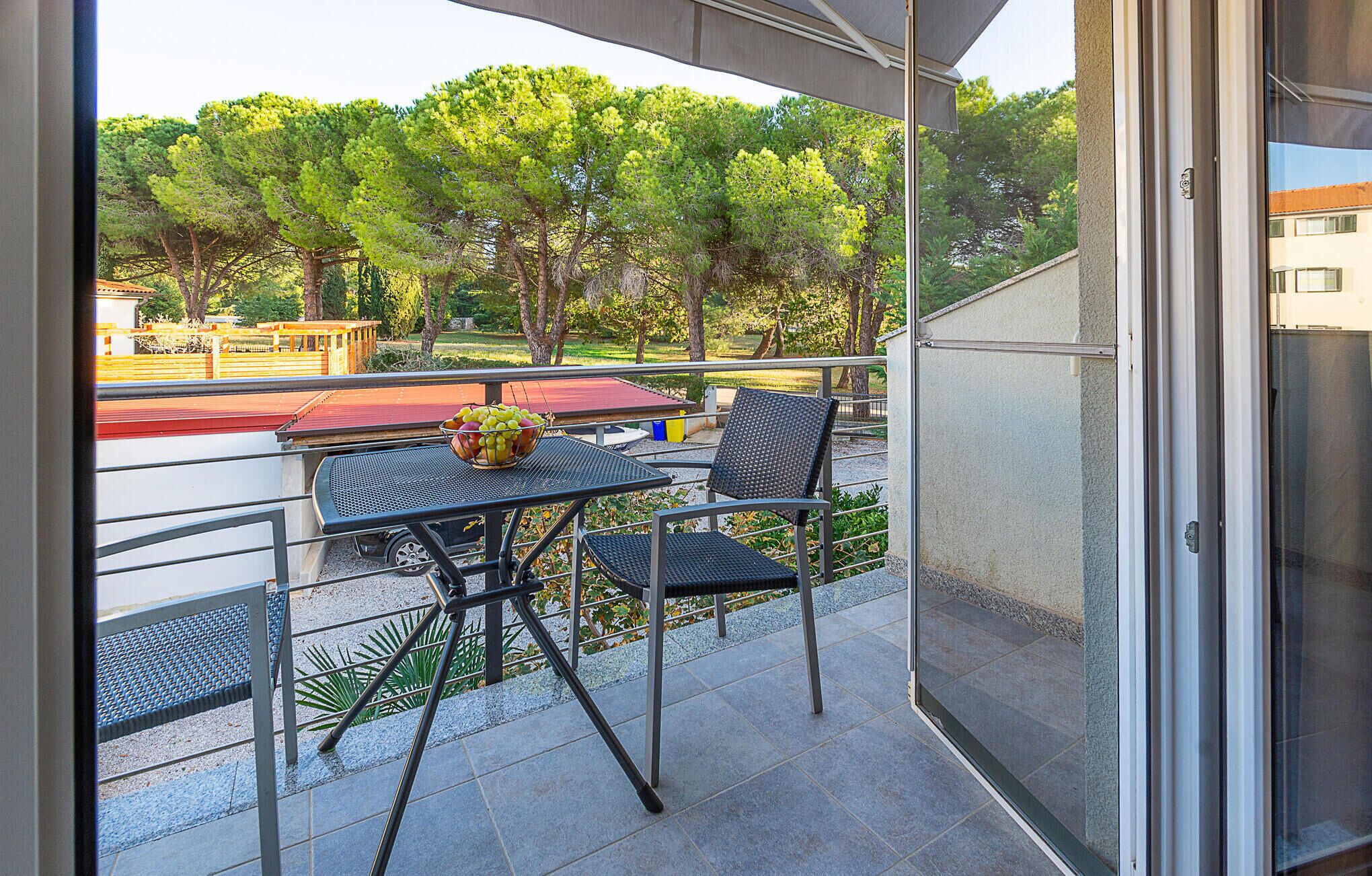 Un balcon avec une table et des chaises, un saladier rempli de fruits et une vue sur les arbres et les bâtiments.