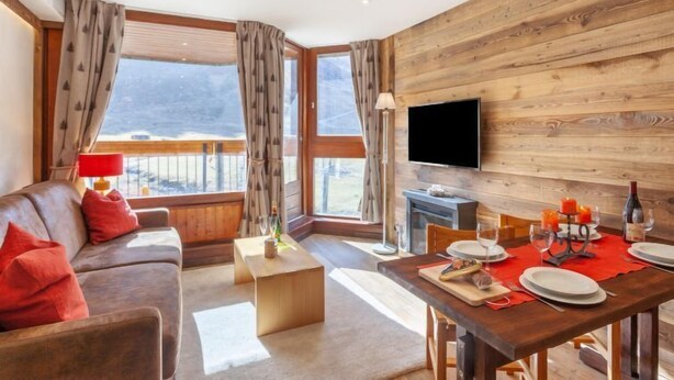 A modern living room with a wooden wall, a sofa with red cushions, a dining table set for two, and a TV mounted on the wall. - Le Borsat (Val Claret) by Interhome (Tignes)
