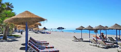 A beach with rows of sun loungers and thatched umbrellas, palm trees, and a clear blue sky.