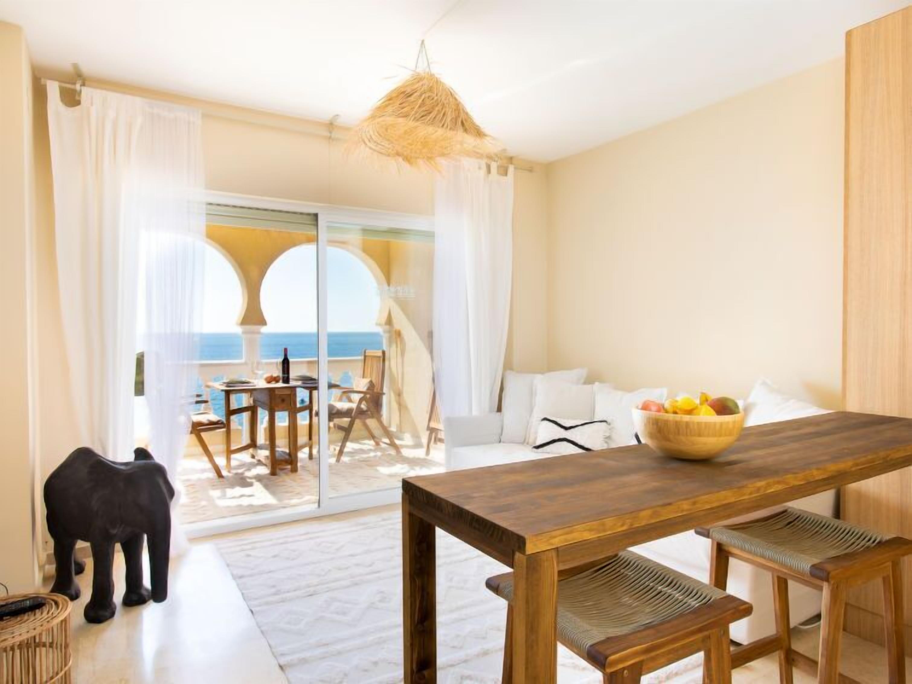 Une pièce avec une table à manger en bois, un saladier rempli de fruits et une vue sur la mer par une porte vitrée.