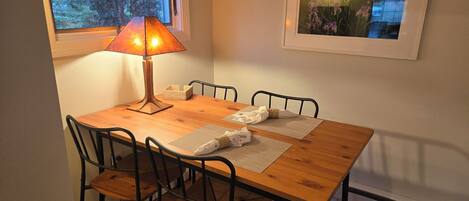 Un comedor con mesa de madera, sillas de metal negro, lámpara de mesa, un cuadro en la pared y ventana con vista a árboles.