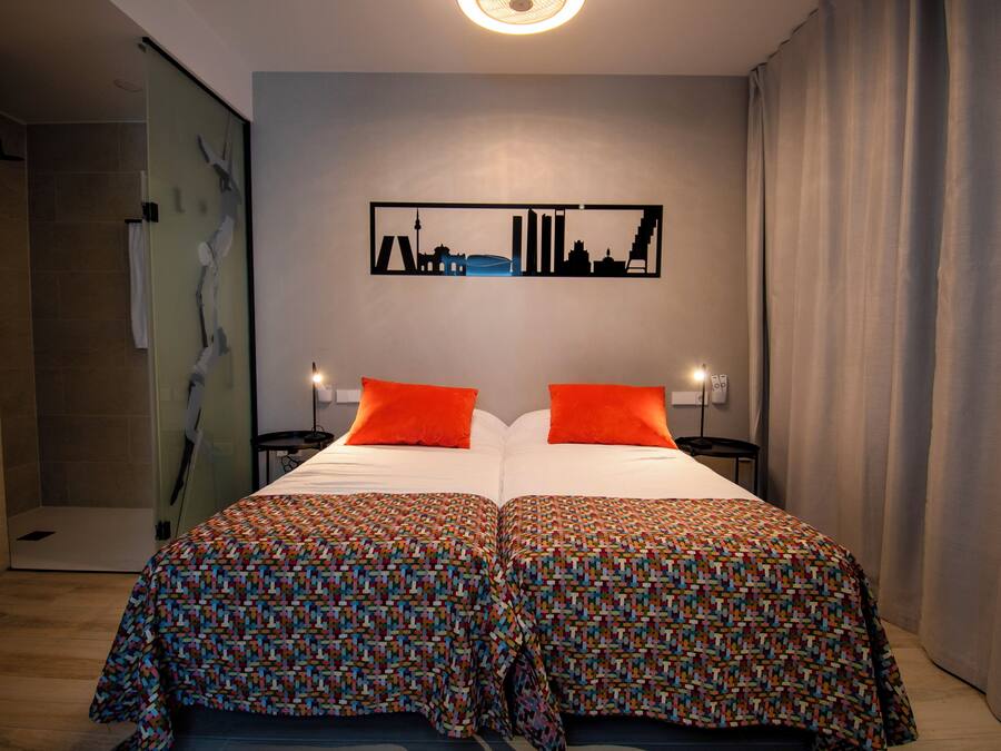 Una cama doble con dos almohadas naranjas, una colcha estampada y una obra de arte con motivo de paisaje urbano en la pared.