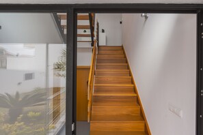 A modern interior with a wooden staircase leading to a landing with a white wall and a small plant. - Secret Beach House (Água de Alto)