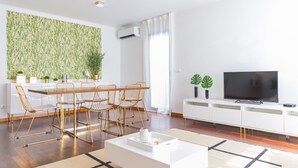 Un salon moderne avec un coin repas, comprenant un grand téléviseur à écran plat, des meubles blancs et un mur décoratif orné d’un motif végétal.