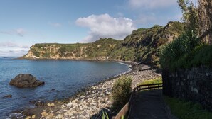 Une route côtière bordée d’un mur de pierres et d’une clôture en bois, qui mène à une plage rocheuse d’où l’on a une vue dégagée sur la mer et les falaises lointaines.