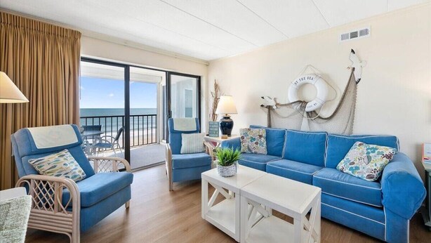 Een kamer met een blauwe bank, twee fauteuils, een witte salontafel en uitzicht op zee door een glazen schuifdeur.
