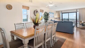 Ein Essbereich mit Holztisch und weißen Stühlen, ein Wohnzimmer mit blauem Sofa und große Fenster mit Blick.
