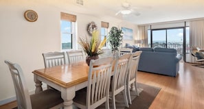 Un comedor con una mesa de madera y sillas blancas, una sala de estar con un sofá azul y ventanas amplias que ofrecen una vista.