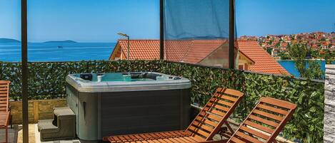 Een balkon met een jacuzzi, twee houten lounge stoelen en uitzicht op zee en gebouwen.