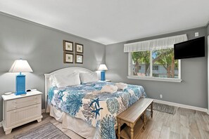 A bedroom with a bed, two bedside tables with lamps, a bench, a window with blinds, and a flat-screen TV mounted on the wall. - Beachside Luxury Villa w/ Pool on Hilton Head (Hilton Head Island)
