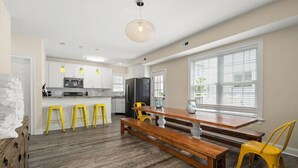 A modern kitchen with white cabinets, a central island, and a dining area with wooden benches and yellow chairs.