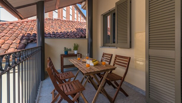 Un espacio al aire libre cubierto con mesa y sillas, una planta en maceta y vistas a un edificio con tejado de tejas rojas.