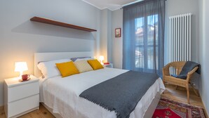 Un dormitorio ordenado con cama, mesitas de noche, una silla y una ventana con cortinas.