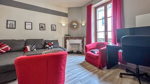 A living room with a grey sofa, red armchair, and a black office chair. There are framed pictures on the wall, a fireplace, and a window with red curtains.