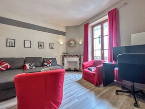 Una sala de estar con un sofá gris, un sillón rojo y una silla de oficina negra. Se observan cuadros enmarcados en la pared, una chimenea y una ventana con cortinas rojas.