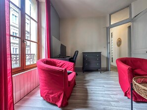 A room with two red chairs, a black TV on a stand, a black dresser, and a window with a view of buildings.