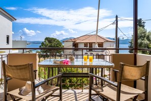 Een balkon met een tafel en stoelen, een boek, twee glazen jus d'orange, en uitzicht op zee en bergen.