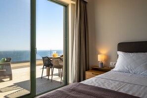 Habitación de hotel con cama, mesita de noche, lámpara, silla, una mesa con una bebida y vista al océano a través de un ventanal.