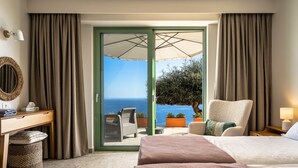 Ein Hotelzimmer mit Bett, Schreibtisch, Stuhl, kleinem Tisch, Lampe und Meerblick durch eine Glastür.