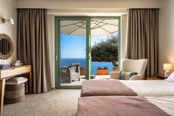 A hotel room with a bed, a desk, a chair, a small table, a lamp, and a view of the sea through a glass door.