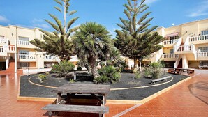 A courtyard with a wooden bench, palm trees, and a building with balconies. - Vacation Home "Sol y Mar Caleta de Fuste" with Shared Pool, Garden & Wi-Fi (Caleta de Fuste)